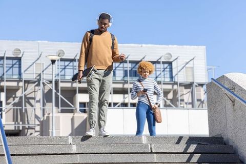 Diverse students descending campus steps carrying skateboard and wearing headphones on sunny day
