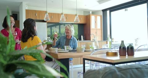 Mature women raising wine glasses laughing around modern wooden kitchen dining table