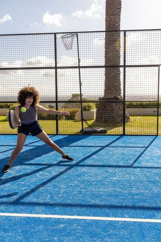 African american woman lunging to hit yellow ball on blue outdoor paddle court in sunlight