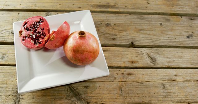 A whole and a partially sliced pomegranate rest on a white square plate, set against a rustic wooden table background, with copy space. Pomegranates are known for their juicy seeds and are often associated with health and vitality.