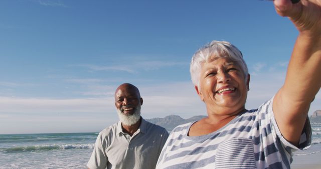 Smiling senior african american couple walking with bicycles and taking a selfie at the beach. healthy outdoor leisure time by the sea.