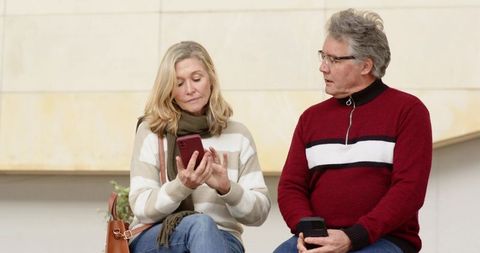 Senior couple comparing smartphones and sharing moment while sitting on bench in urban courtyard