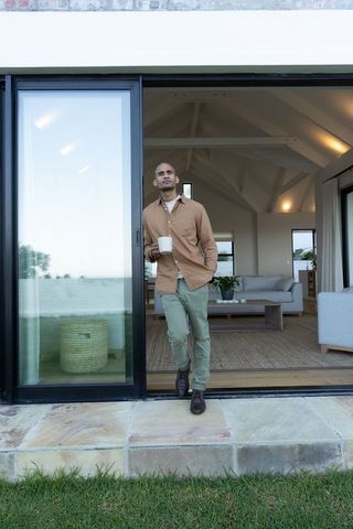Man leaning in sliding glass doorway holding coffee mug overlooking lawn and living room