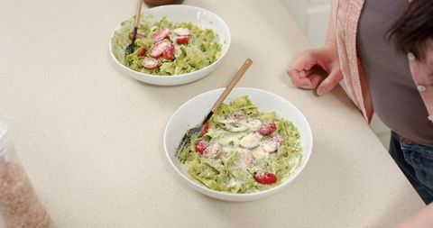 Asian woman preparing pesto pasta with cherry tomatoes and parmesan on kitchen counter