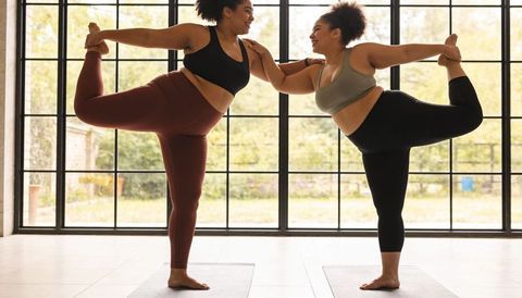 Partner yoga practicing dancer pose on mats by floor-to-ceiling window with garden view