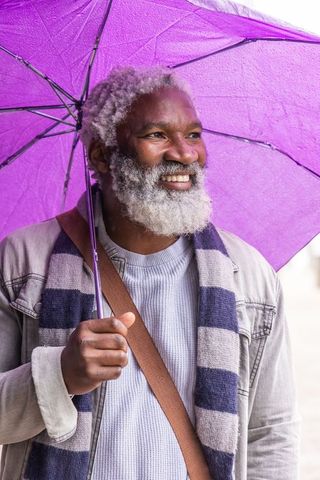 Senior african american man smiling under purple umbrella on rainy urban street