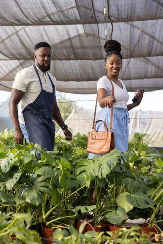 Woman pointing at monstera plants while nursery worker assisting under shade canopy