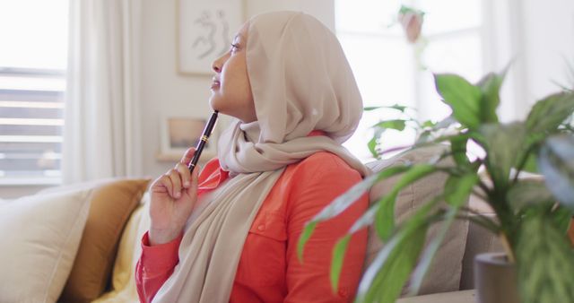 Image of thoughtful biracial woman in hijab holding pen and writing in living room at home. Happiness, creativity, relaxation, inclusivity and domestic life.