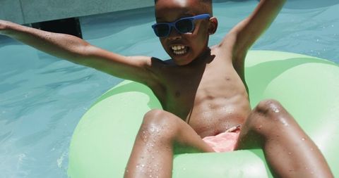 African american boy floating on green pool tube wearing blue sunglasses enjoying summer