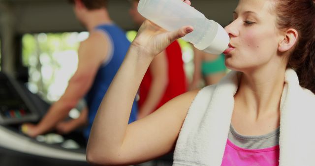Young woman with towel around neck and water bottle hydrating after workout session in gym. Use for fitness advertising, health promotions, gym membership marketing, and hydration awareness campaigns.