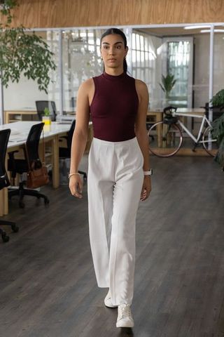 Confident young woman walking in modern open-plan office wearing burgundy top and white trousers