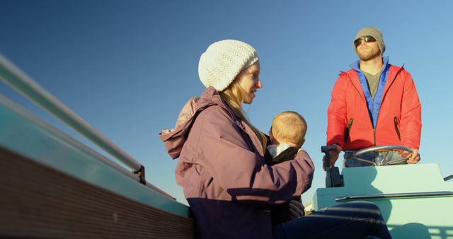 Capture of a family enjoying a boat ride on a sunny day. Mother holding child while father navigates the boat. They are all dressed in colorful clothing, indicative of cool weather. Ideal for use in content related to family bonding, outdoor activities, leisure time, and warm spring days.