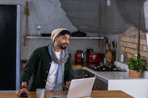 Bearded man working from home at kitchen island with laptop and mug, cozy rustic workspace