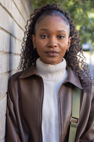 Confident young woman standing by brick wall wearing brown leather jacket and turtleneck