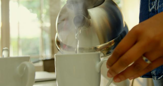 Close-up of a hand pouring hot water from a kettle into a white mug, with steam visible, set in a bright, sunlit kitchen. Ideal for use in articles or advertisements about morning routines, home cooking, or coffee and tea products.