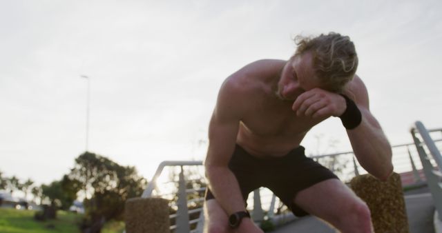 Caucasian shirtless man wiping sweat from his forehead on bridge in city. Sports, fitness, healthy living and outdoor activities, unaltered.