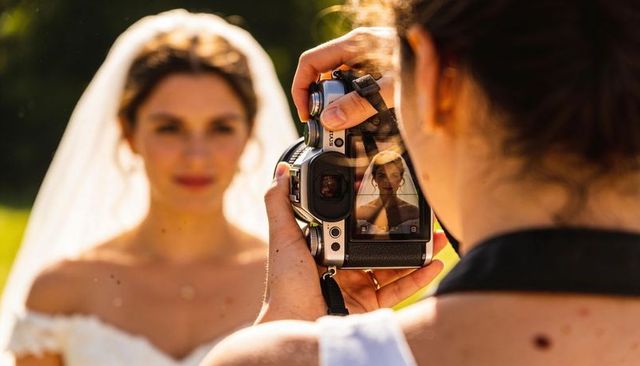Wedding photographer framing bride through dslr viewfinder veil sunlit garden portrait
