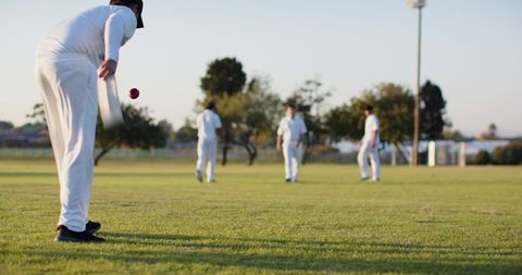 Cricket bowler bowling red ball during golden hour on grassy outfield with fielders