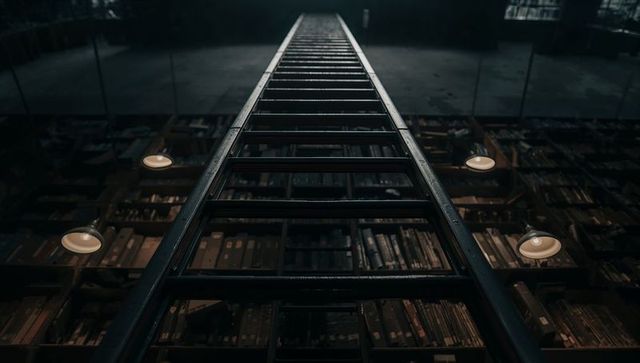 Tall rolling library ladder climbing toward dim mezzanine with vintage lamps and archives