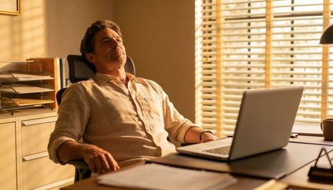 Middle-aged man leaning back at desk gazing upward in sunlit minimalist office with laptop