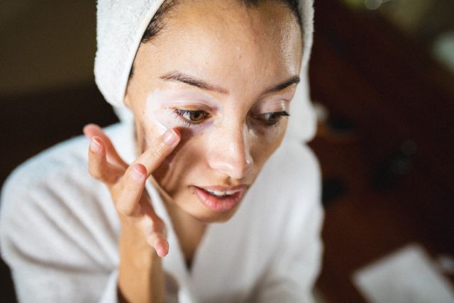 Close-up of biracial young woman in bathrobe applying cream on face during spa - Download Free Stock Photos Pikwizard.com