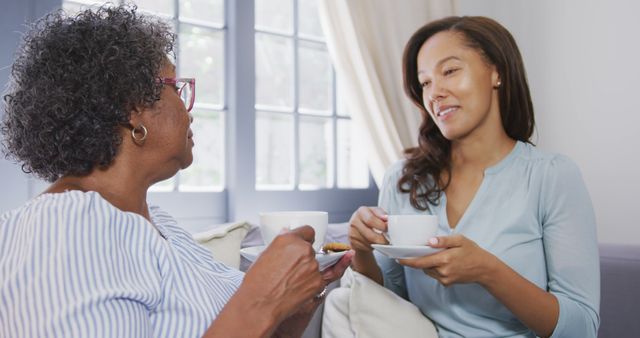 Happy senior african american woman and her adult daughter sitting on sofa, drinking tea and talking. Lifestyle, free time, family and domestic life.