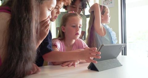 Multiethnic children collaborating on tablet in bright classroom leaning around screen
