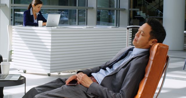 Middle-aged businessman in suit sleeping in office lounge, taking a break from work. Modern office environment with glass walls and another employee working in background. Useful for themes related to workplace stress, corporate life balance, productivity, burnout, or office interior design.