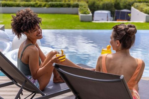Two diverse women relaxing poolside on lounge chairs holding orange cocktails and laughing