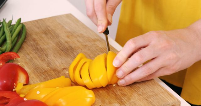 Hands are slicing a yellow bell pepper on a wooden cutting board. Fresh vegetables like red bell pepper and green beans surround the cutting area. Ideal for use in cooking blogs, healthy eating articles, and meal prep tutorials.