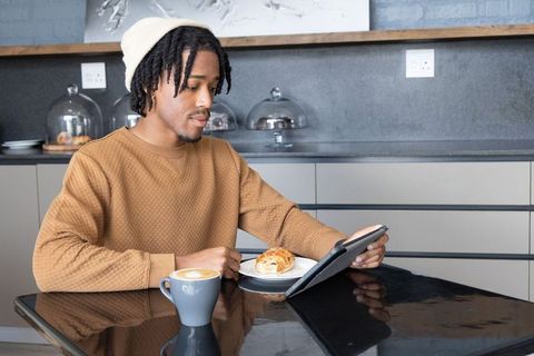 African american man reading tablet while enjoying coffee and croissant in modern kitchen