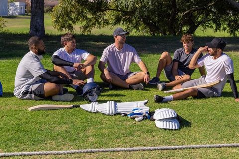 Cricket teammates resting and chatting on grass after practice, holding bats and gear