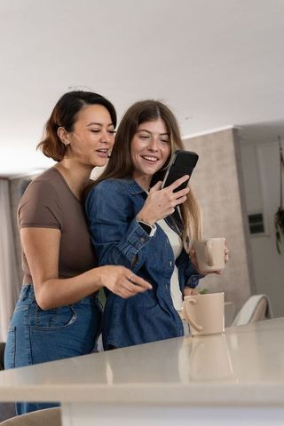 Diverse female couple sharing coffee and smartphone moment in modern kitchen