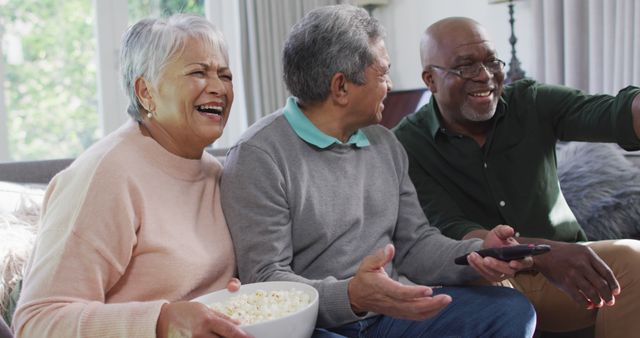 Three senior friends are sitting on a couch in the living room, sharing a bowl of popcorn and enjoying a TV show together. They are laughing, and one holds a remote control while another points at the television. This scene could be used to illustrate friendship, leisure, happiness, and companionship among elderly individuals. It's great for promoting senior living communities, social engagement activities, or any content focused on positive aging and senior well-being.