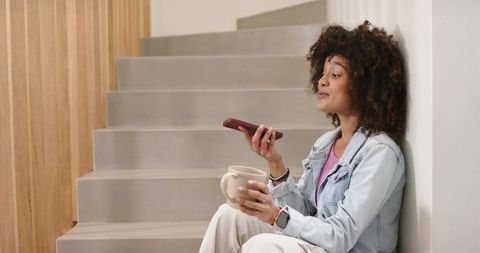 Young woman sitting on modern stairs holding smartphone and coffee mug, speaking