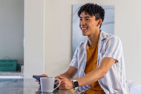 Young man smiling at kitchen counter holding smartphone and mug, wearing smartwatch