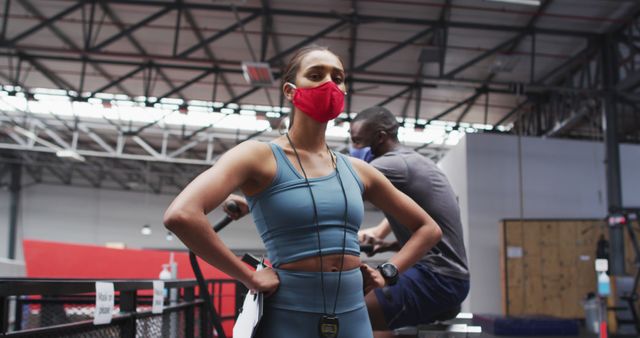 Female fitness trainer wearing a red mask oversees a male athlete working out on exercise machines in a spacious, modern gym. The gym environment features structural beams and open space fostering an atmosphere for various types of exercise. This image is excellent for illustrating fitness training during health precautions, promoting healthy living even during pandemics, personal training services, or fitness facility advertisements.