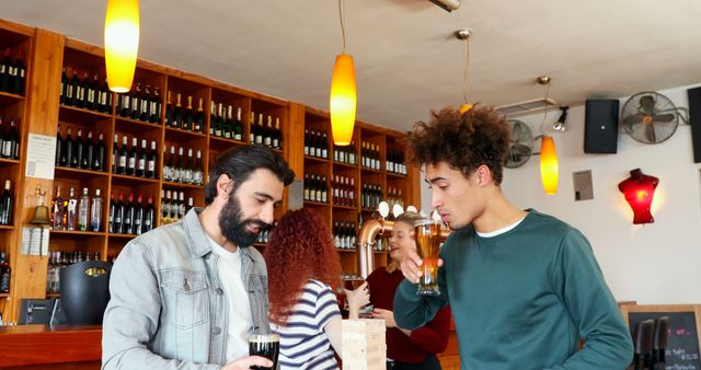 Group of friends seen socializing and enjoying drinks at a modern bar while playing a game of Jenga. Young adults are engaged in conversation and leisure activities. This scene is ideal for illustrating social gatherings, nightlife, or casual fun settings.