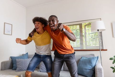 Cheering diverse male friends celebrating sports win while leaning over sofa at home
