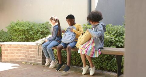 Schoolchildren sitting and unpacking backpacks on bench in sunlit campus courtyard