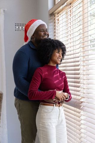 African american couple embracing by window wearing santa hat sharing cozy holiday moment