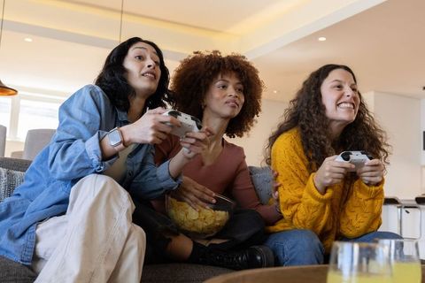 Laughing diverse women playing video games on sofa holding white controllers and snacking