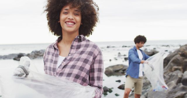 Portrait of african american woman smiling while collecting garbage on the rocks near the sea. environmental protection and recycling concept