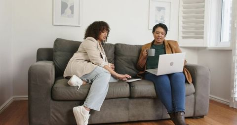 Two female colleagues collaborating on laptop while sitting on gray sofa in living room
