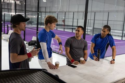 Padel team camaraderie: multicultural athletes chatting over coffee beside indoor court