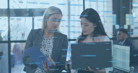 Female colleagues reviewing analytics on laptop and tablet, collaborating in modern office