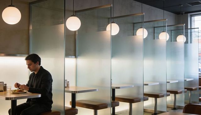 Solitary man sitting and eating at modern glass-partitioned cafe booth with globe lights