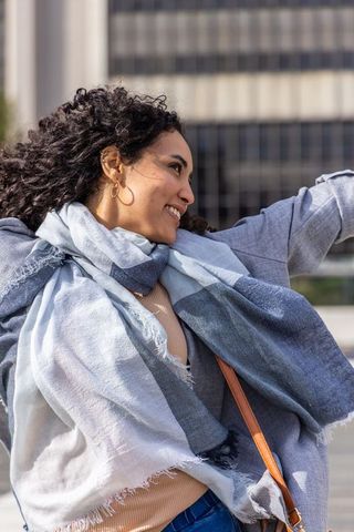 Smiling woman enjoying breezy urban plaza wearing oversized scarf, gray coat, denim
