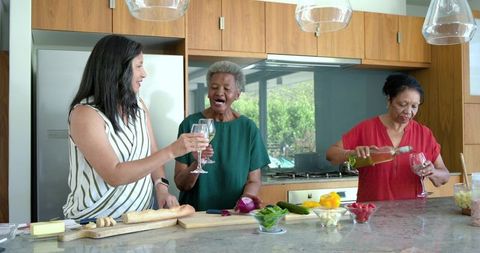 Senior women toasting, laughing and preparing food at kitchen island while pouring wine