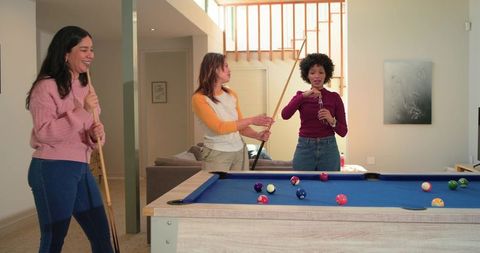 Diverse female friends laughing and playing pool while opening bottle in modern lounge